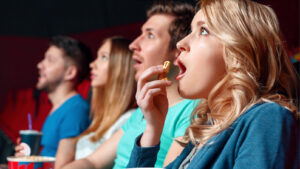 a row of people sitting in a movie theatre eating popcorn and looking in awe at a promo video