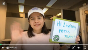 A screenshot of a Vidyard video featuring a female salesperson waving at the camera while holding a small sign that says "Hi Lindsey, press play."