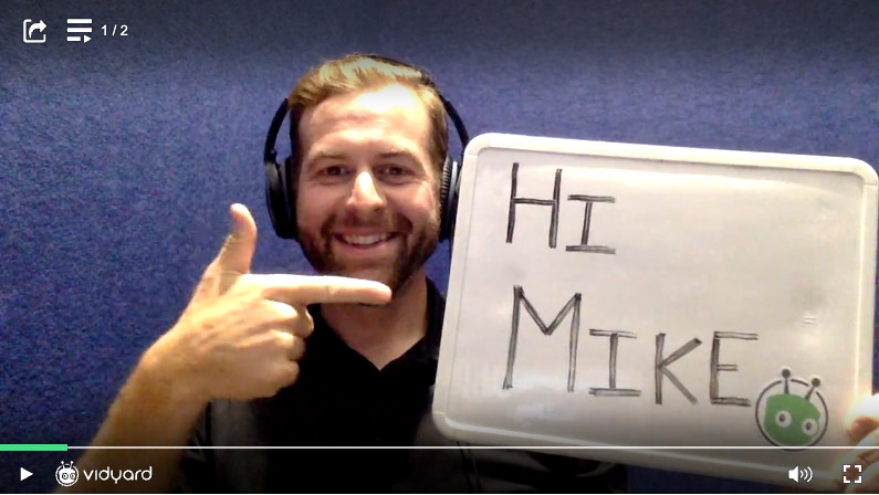A screenshot of a Vidyard video featuring a male salesperson holding up a small sign that reads "Hi Mike"