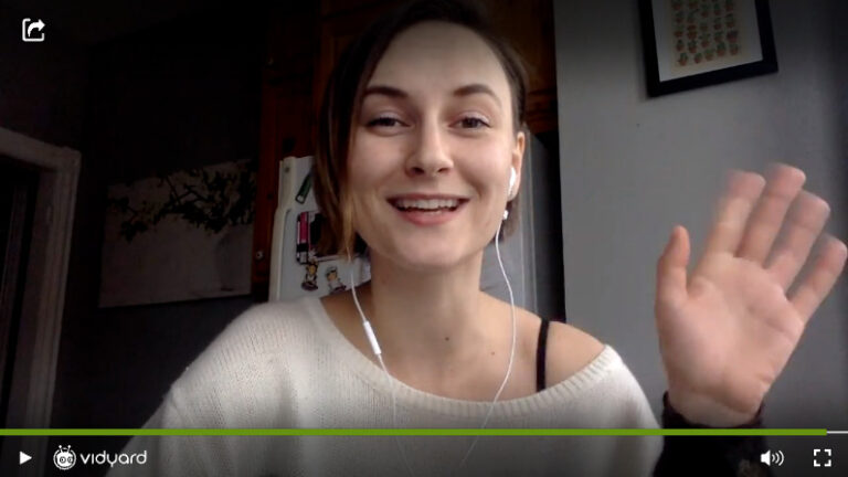 A screenshot of a Vidyard video featuring a female salesperson smiling and waving at the camera